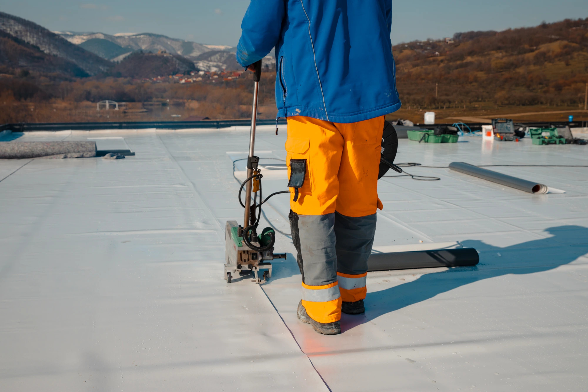  Roofer using seam welding equipment on a white TPO membrane flat roof with rolled membrane material nearby